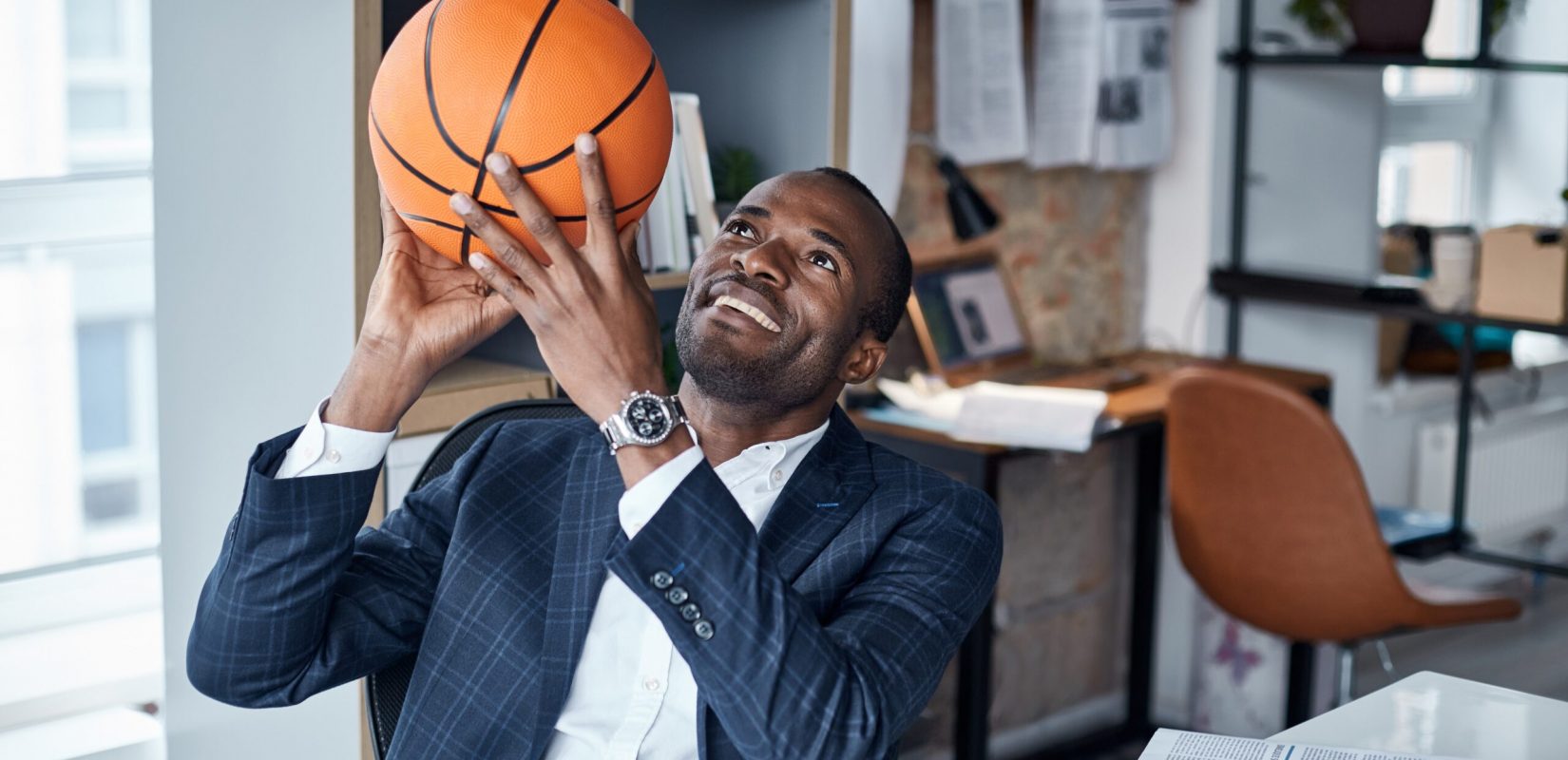 Playful mood. Cheerful young african manager in suit is sitting in office and holding orange basketball with smile. He is looking up while being ready to throwing it